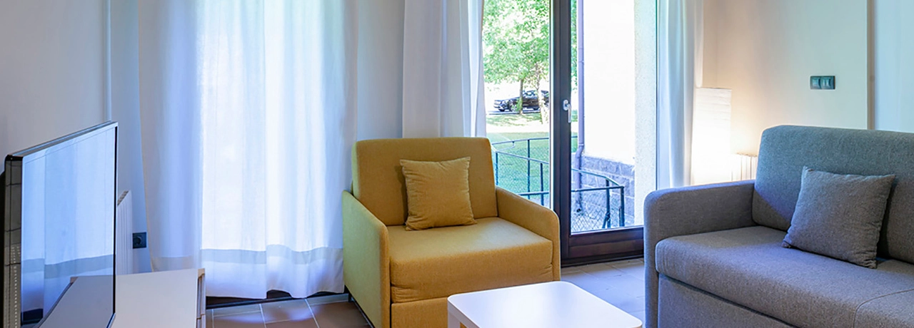 Sala luminosa con sill&oacute;n amarillo, sof&aacute; gris y mesa blanca frente a una ventana.