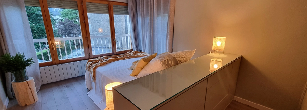 Dormitorio iluminado con cama, lámparas y ventana grande al fondo.