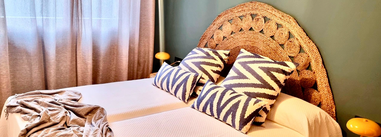 Cama con cojines geométricos y cabecero de mimbre, cortinas y lámparas amarillas.