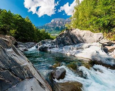 R&iacute;o cristalino entre rocas y &aacute;rboles con monta&ntilde;as al fondo bajo cielo azul.