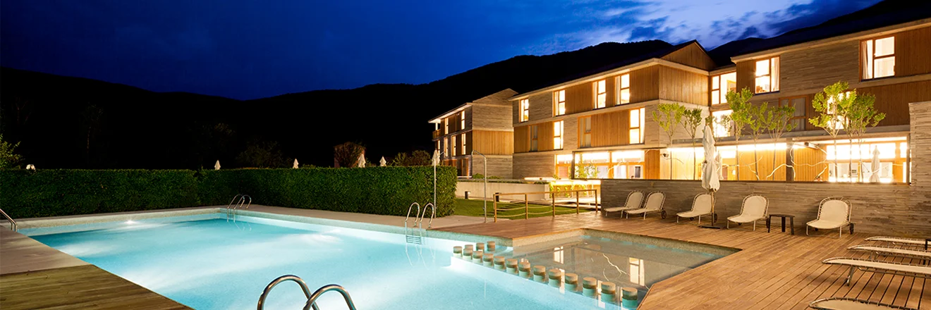 Piscina iluminada frente a un hotel moderno al anochecer.