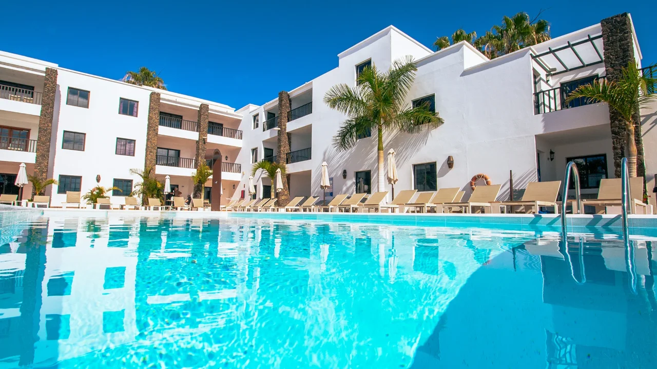 Piscina frente a un edificio blanco con palmeras y sillas reclinables.