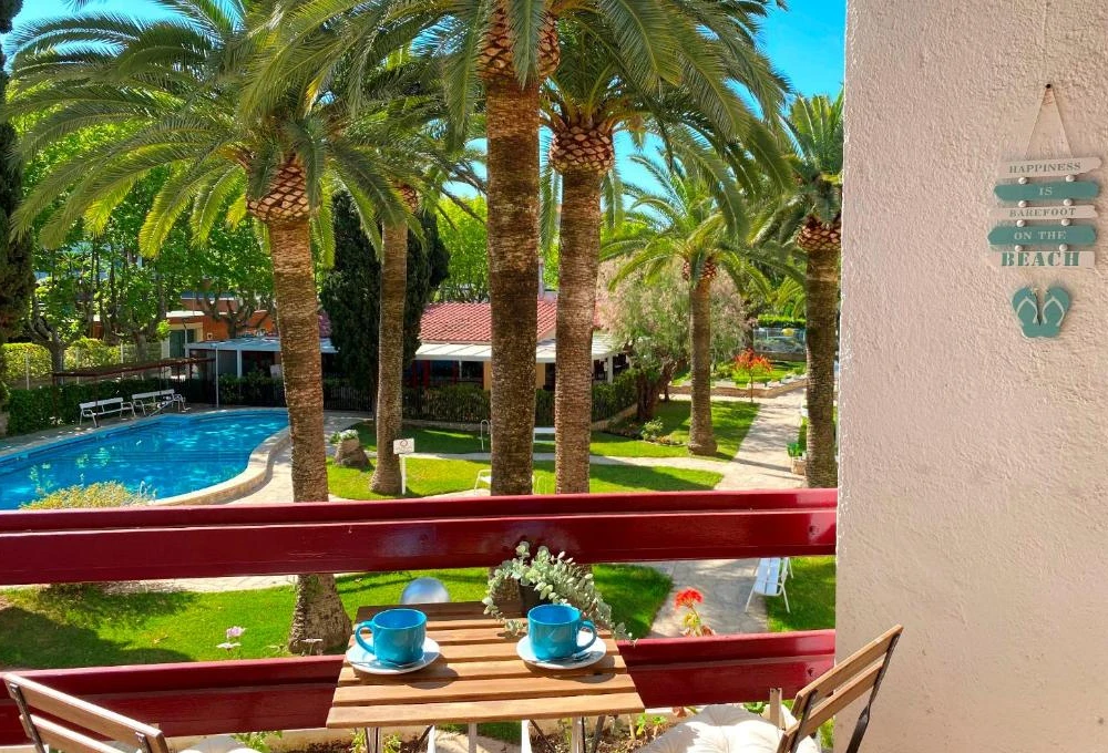 Balc&oacute;n con vista a piscina y palmeras, mesa con tazas. Cartel decorativo.