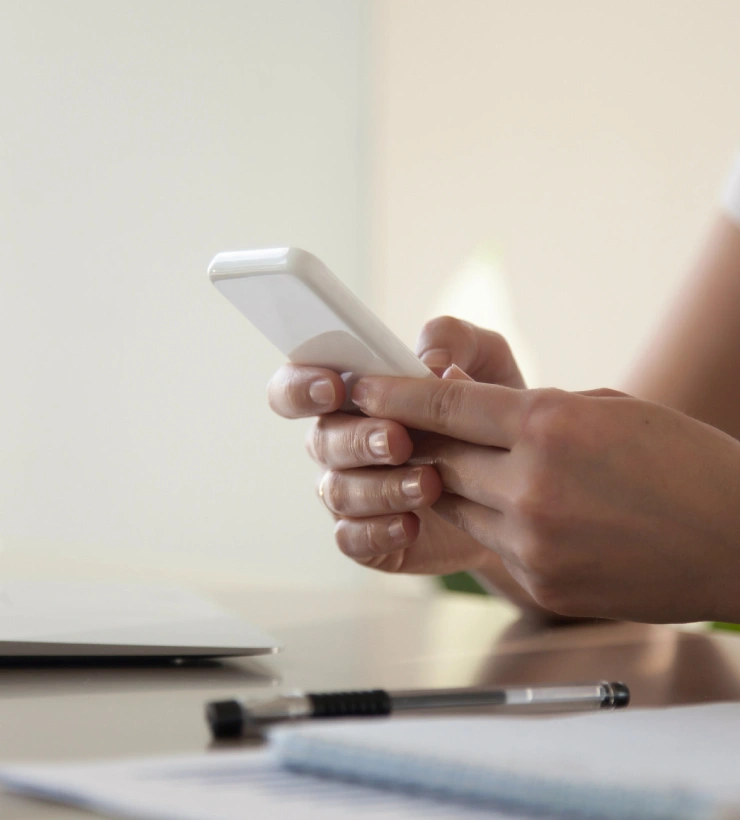 Persona usando un celular junto a un bol&iacute;grafo y papeles sobre la mesa.