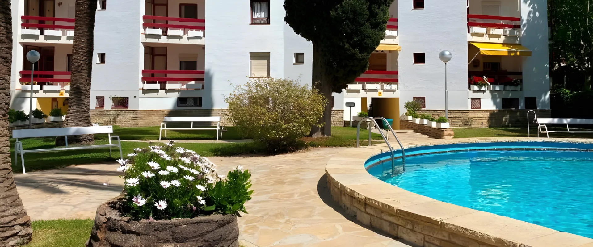 Piscina en un jard&iacute;n junto a edificio blanco con balcones rojos y flores.