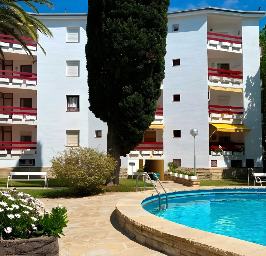 Edificio blanco con balcones rojos y piscina rodeada de jard&iacute;n.