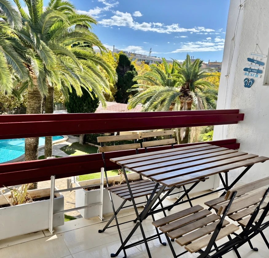 Balc&oacute;n con mesa y sillas de madera, vista de palmeras y piscina al fondo.