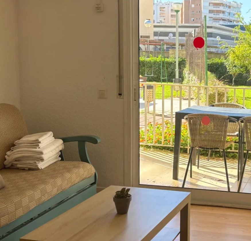 Sala con sof&aacute;, mesa, jard&iacute;n exterior y edificios al fondo.