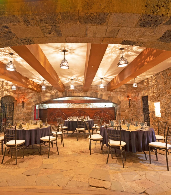 Sala de eventos r&uacute;stica con mesas redondas y sillas, techo con vigas y l&aacute;mparas.