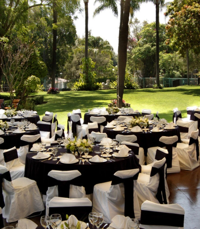 Mesas elegantes al aire libre con manteles negro y blanco, rodeadas de vegetaci&oacute;n.