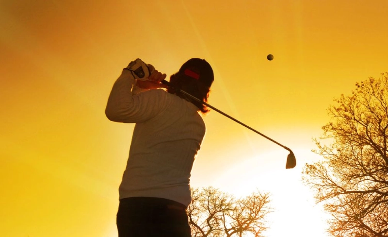 Persona jugando golf al atardecer, con la pelota en el aire.