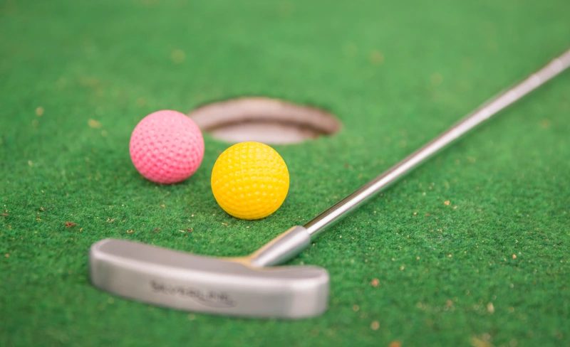 Palos y bolas de mini golf, una amarilla y una rosa, sobre c&eacute;sped verde.