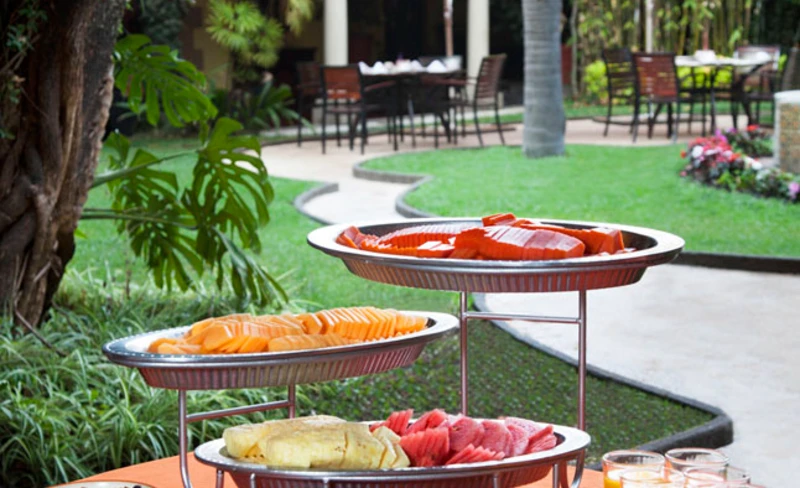 Mesa con frutas y asados en parrilla sobre c&eacute;sped, en jard&iacute;n tropical