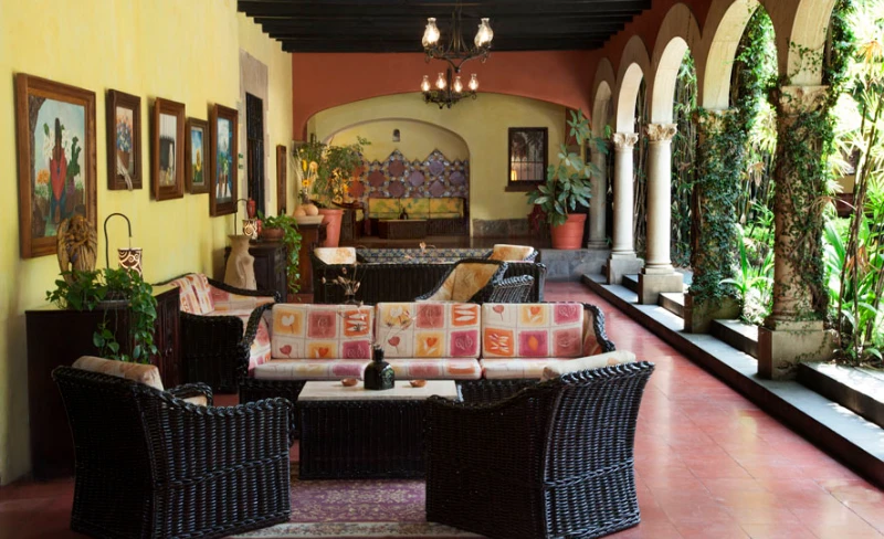 Terraza con sof&aacute; y sillones de mimbre, columnas y jard&iacute;n con plantas.