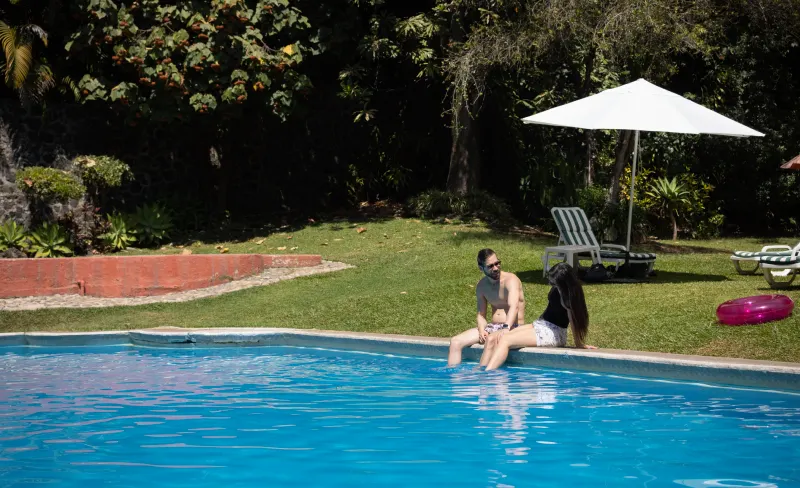 Persona en la orilla de una piscina bajo una sombrilla blanca en un jard&iacute;n.