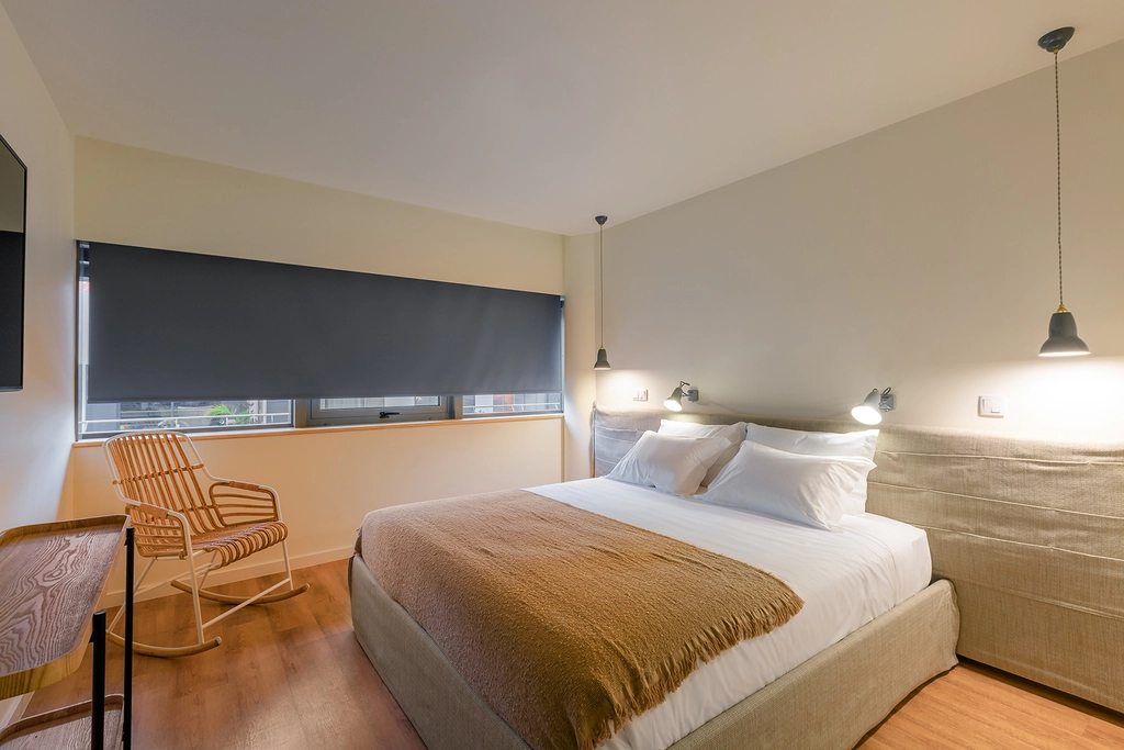 Quarto moderno com cama de casal, cadeira de madeira, lumin&aacute;rias e cortina cinza.