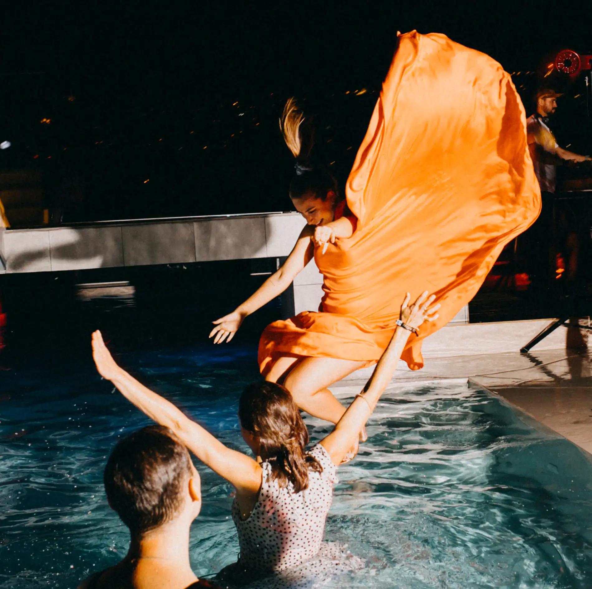 Mulher em vestido laranja pulando em piscina &agrave; noite, pessoas aguardam na &aacute;gua.