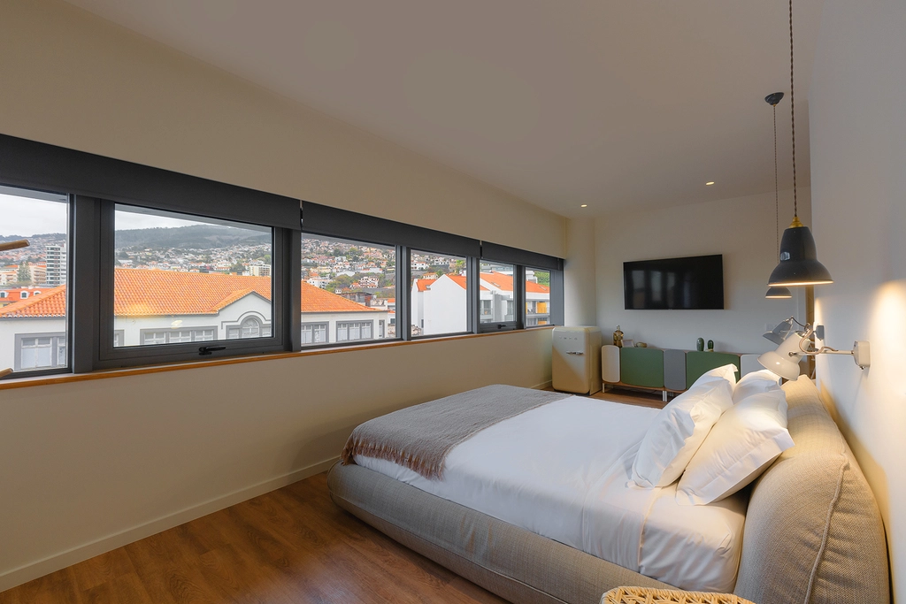 Quarto moderno com cama de casal, TV, decora&ccedil;&atilde;o minimalista e janelas com vista panor&acirc;mica da cidade.