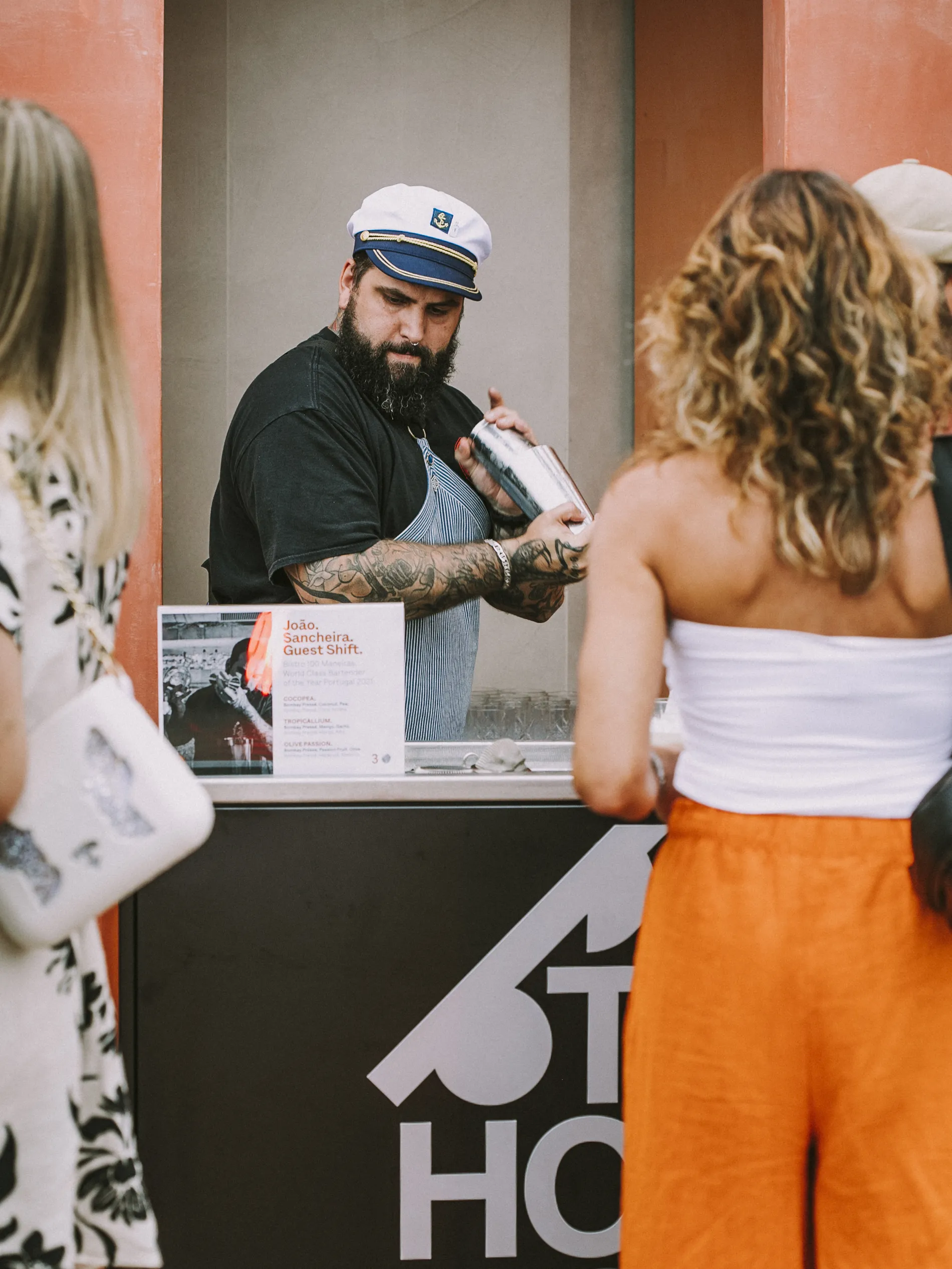 Bartender de chap&eacute;u naval prepara drinque em um quiosque para clientes esperando.