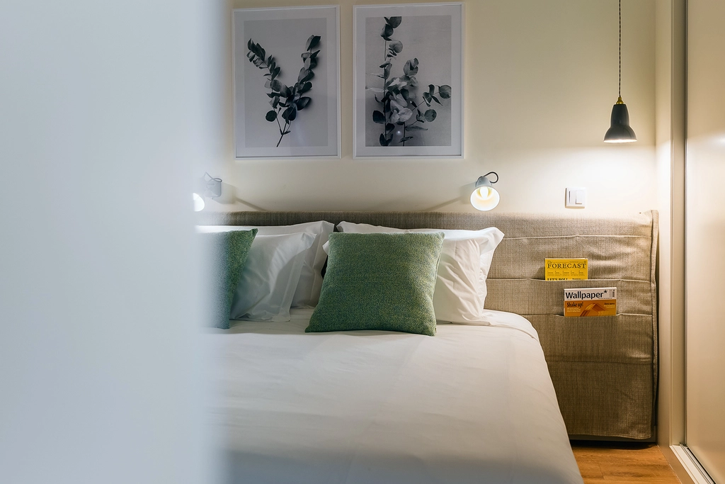 Quarto moderno com cama, almofadas verdes e abajur, decorado com quadros de folhagens.