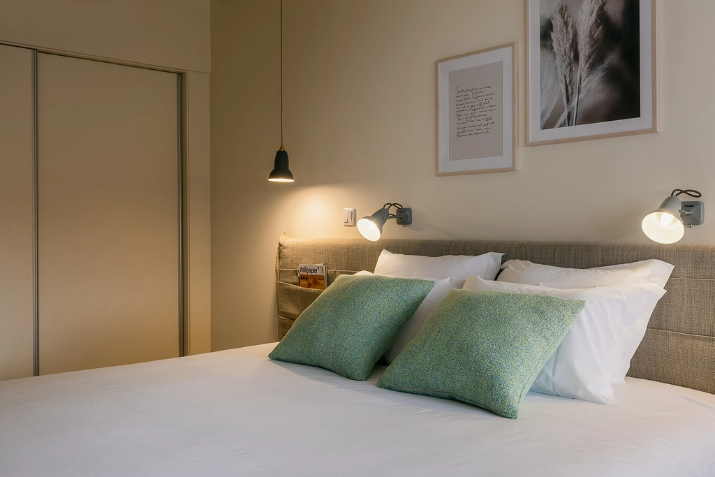 Quarto moderno com cama de casal, almofadas verdes e lumin&aacute;rias nas laterais.