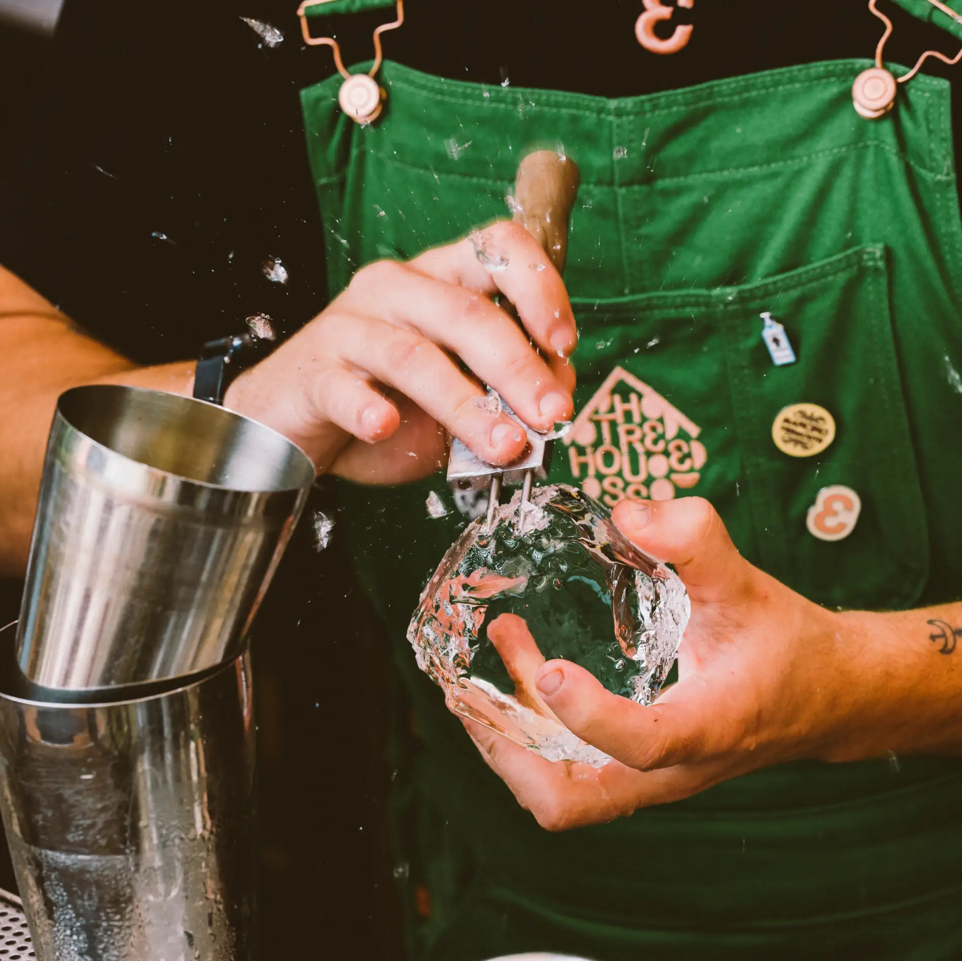 Pessoa esculpindo um bloco de gelo com ferramenta, ao lado de copo de metal em uma bancada.