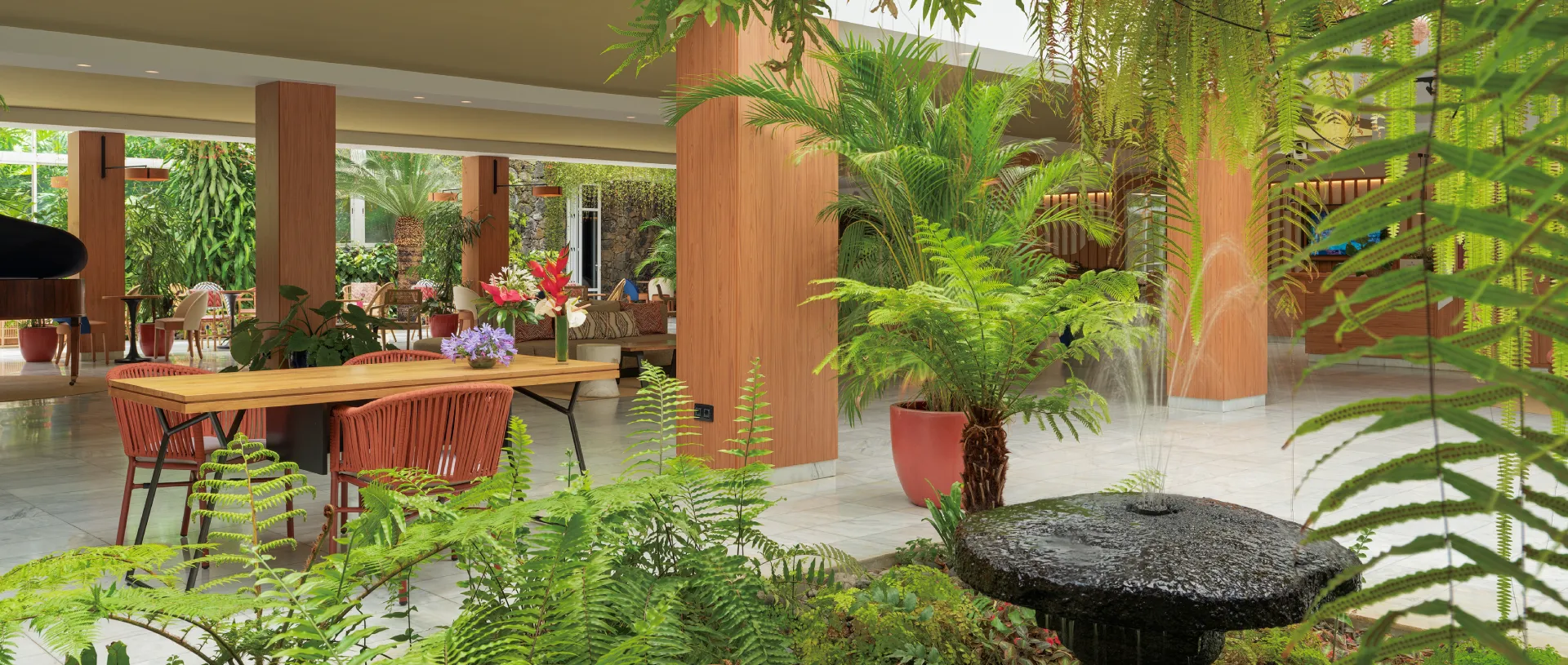&Aacute;rea de descanso con plantas, mesas de madera y fuente de agua en interior.