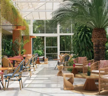 Terraza luminosa con mesas, sillas de mimbre y plantas tropicales.