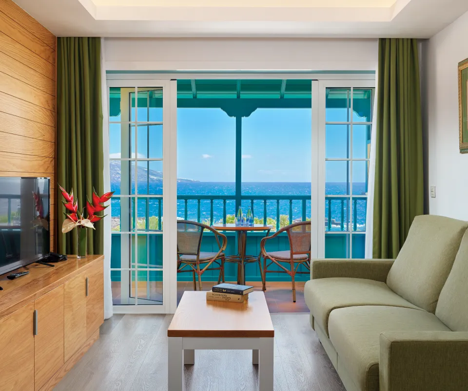 Sala con vista al mar, sof&aacute; verde y mesas de madera.