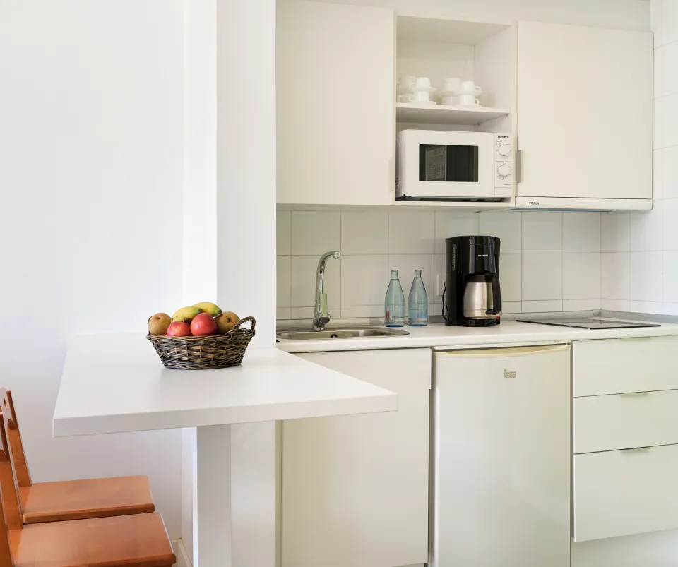 Cocina blanca con mesa, canasta de frutas, microondas y cafetera.