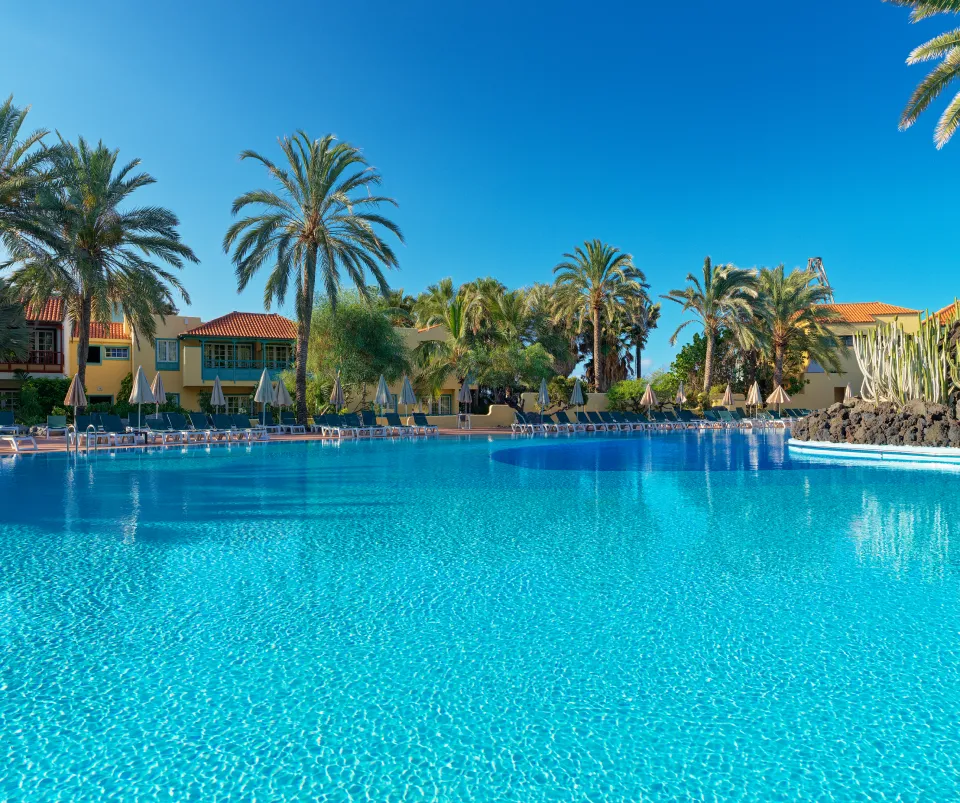 Piscina azul rodeada de palmeras y tumbonas bajo un cielo despejado.