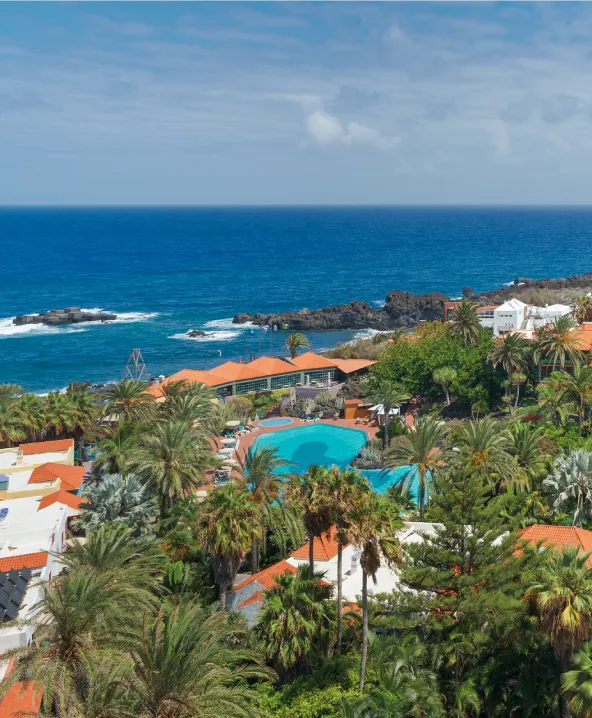 Complejo tur&iacute;stico junto al mar con palmeras y piscina bajo un cielo azul.