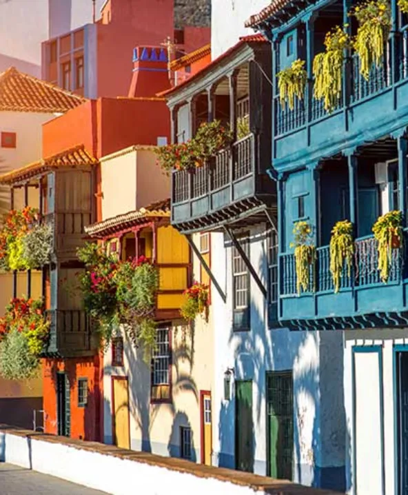 Casas coloridas con balcones y plantas colgantes en una calle iluminada por el sol.