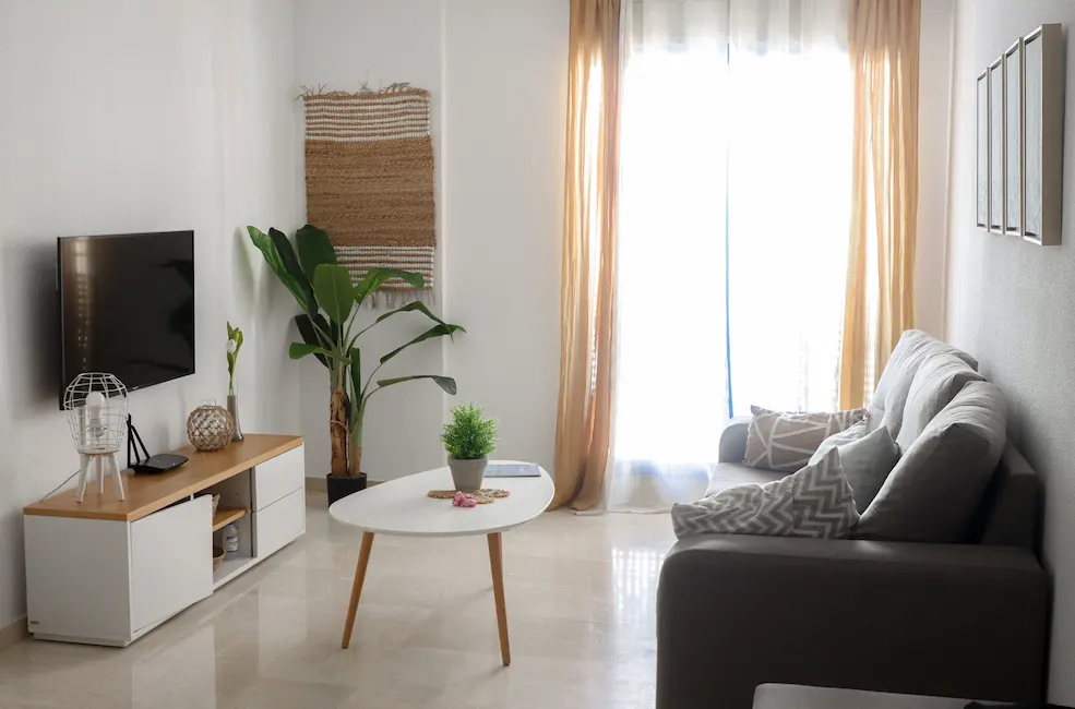 Sala moderna con sofá, mesa y televisor. Ventana con cortinas ligeras.