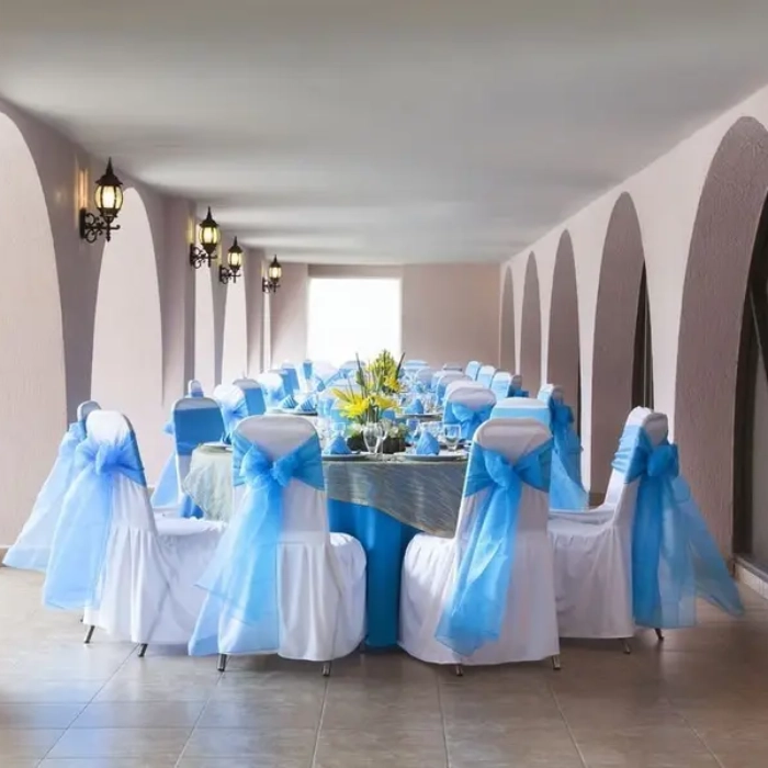 Mesa decorada para evento, sillas con lazos azules bajo arcos iluminados.