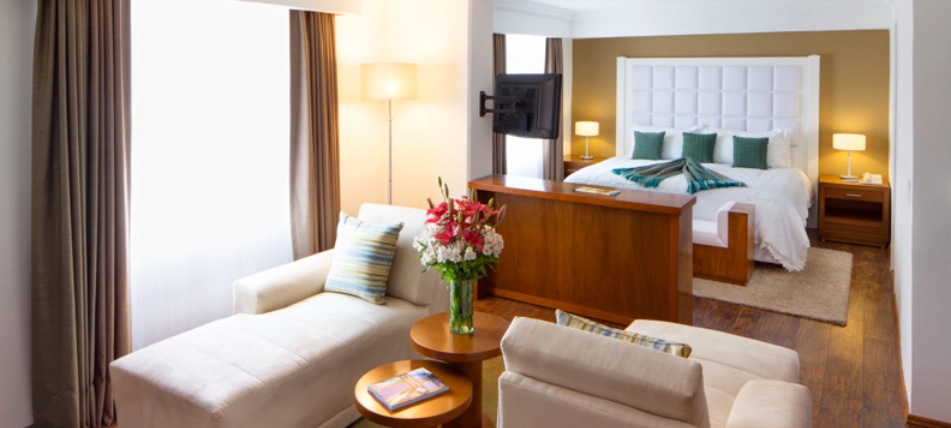 Sala y dormitorio moderno con sof&aacute;, cama, y flores en mesa central.