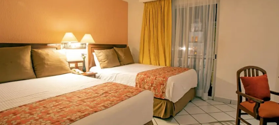 Habitaci&oacute;n de hotel con dos camas, l&aacute;mparas, cortinas y silla.