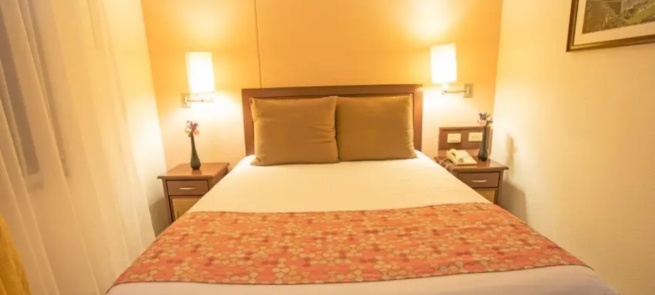 Cama matrimonial en habitaci&oacute;n iluminada con dos l&aacute;mparas y decoraci&oacute;n simple.