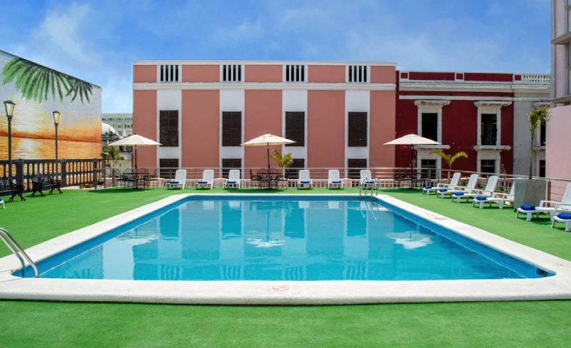 Piscina al aire libre rodeada de sillas y sombrillas, edificio al fondo.