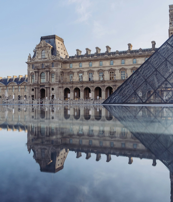 B&acirc;timent historique et pyramide en verre se refl&eacute;tant dans l'eau, ciel d&eacute;gag&eacute;.