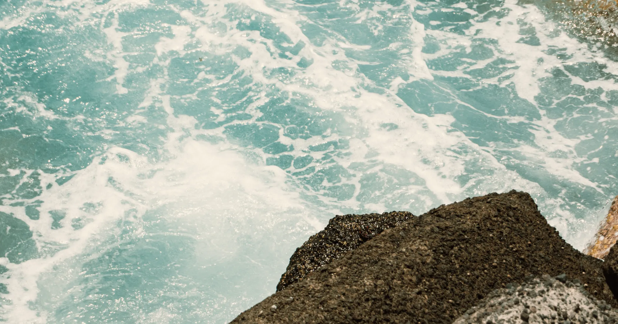 Rocas oscuras junto a agua turquesa con espuma blanca en movimiento.