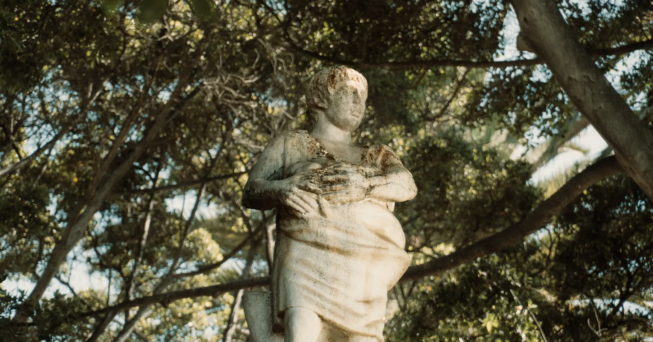 Estatua de m&aacute;rmol antigua entre &aacute;rboles y hojas verdes.