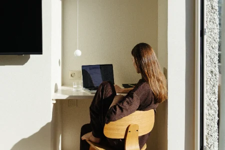 Persona sentada en una silla frente a una computadora port&aacute;til en un rinc&oacute;n iluminado por luz natural.