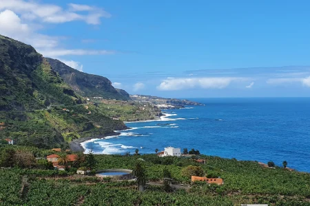 Costa rocosa con monta&ntilde;as y vegetaci&oacute;n junto al mar azul bajo un cielo parcialmente nublado.