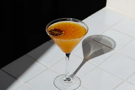 C&oacute;ctel en copa de martini con rodaja de c&iacute;trico sobre mesa blanca.