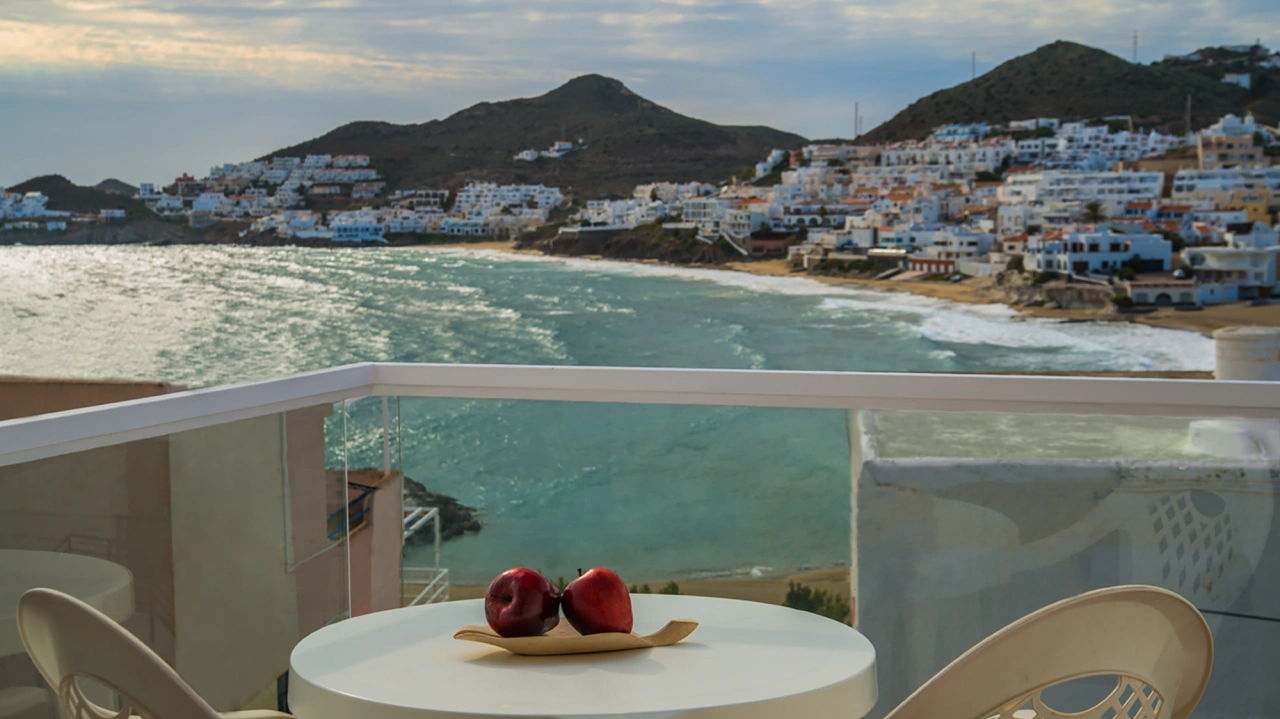 Terraza con manzanas sobre mesa, vista al mar y casas blancas en la costa.