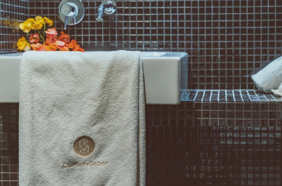 Toalla blanca colgada en lavabo con azulejos oscuros y flores naranjas.