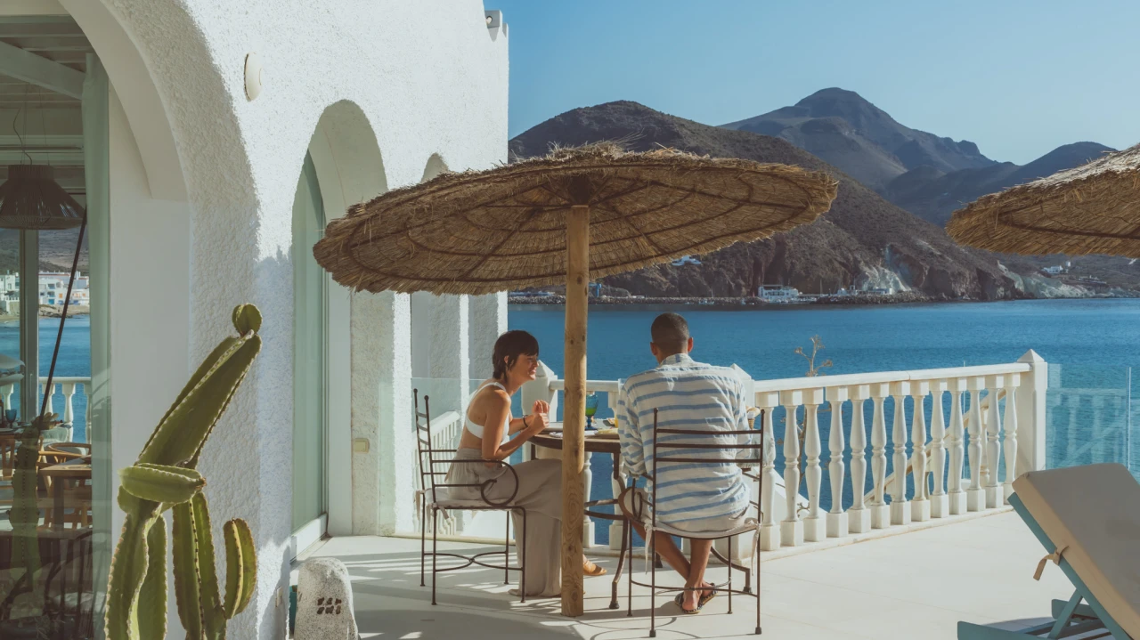 Pareja desayunando en terraza junto al mar, paisaje monta&ntilde;oso al fondo.