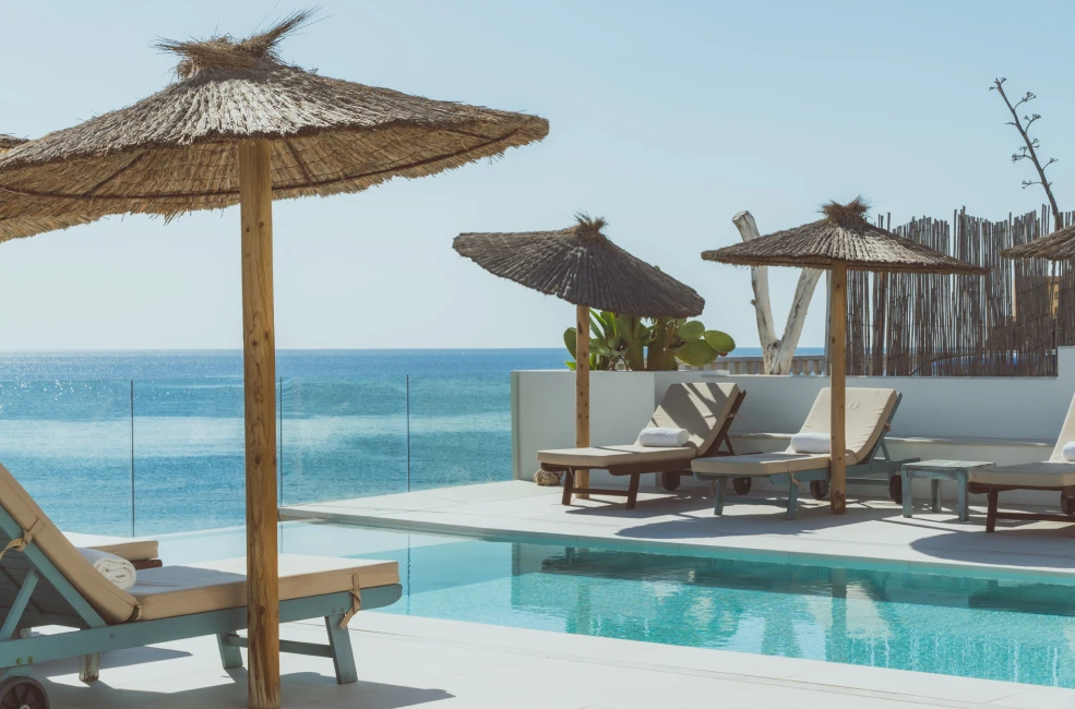 Piscina junto al mar con tumbonas y sombrillas de paja.