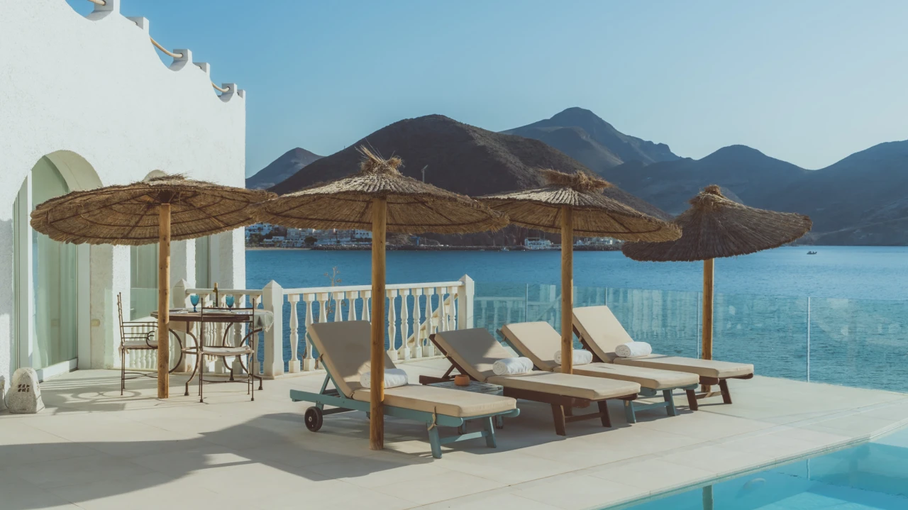 Terraza con tumbonas y sombrillas junto al mar y monta&ntilde;a al fondo.